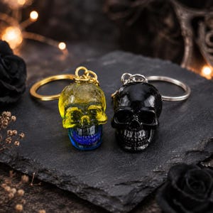 May include: Two skull keychains with metal rings. One skull is translucent yellow with a blue jaw and gold-toned ring. The other skull is solid black with a silver-toned ring. Both feature detailed eye sockets, nasal cavities, and teeth.