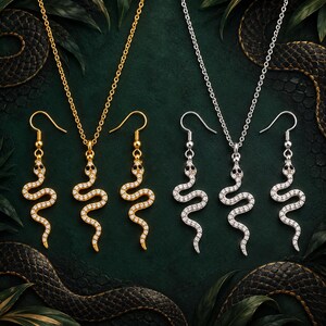 May include: Two jewellery sets featuring snake-shaped pendants and matching drop earrings. One set is gold-toned with clear crystals and black stone eyes, while the other is silver-toned with clear crystals and black stone eyes on thin chains.