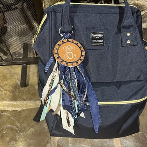 May include: A navy blue backpack with a gold zipper and a brown leather keychain with a monogrammed initial "G" and blue and white ribbons.