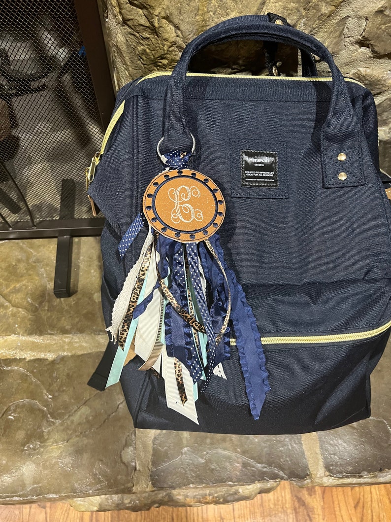 May include: A navy blue backpack with a gold zipper and a brown leather keychain with a monogrammed initial "G" and blue and white ribbons.