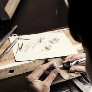 May include: A person working on a silver ring with a small tool. The ring is on a wooden surface and there are other silver rings on a white piece of paper.