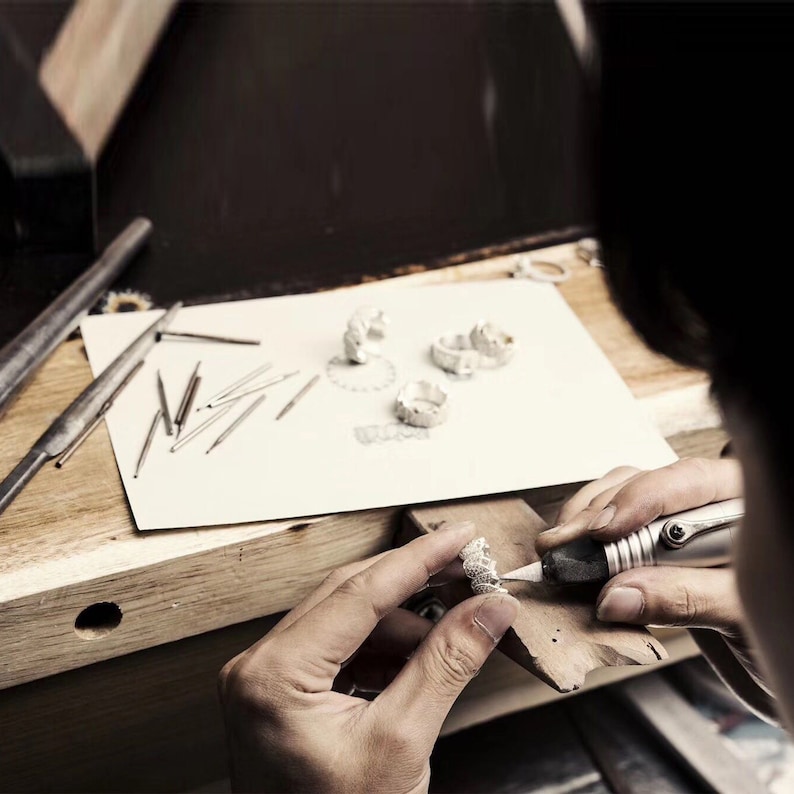 May include: A person working on a silver ring with a small tool. The ring is on a wooden surface and there are other silver rings on a white piece of paper.