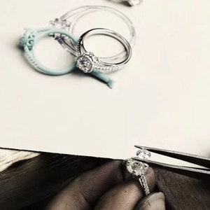 May include: A close-up of a jeweler setting a diamond in a silver ring. The ring is being held in place with tweezers.