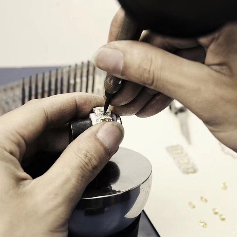 May include: A close-up of a jeweler using a tool to set a small, square-shaped diamond in a piece of jewelry. The jewelry is being held in place by a metal clamp.