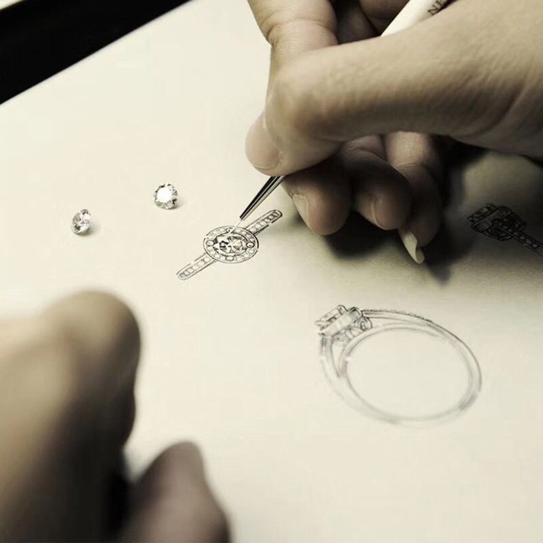 May include: A hand sketches designs for engagement rings on a piece of paper. The rings are drawn in pencil and feature different styles of diamonds.
