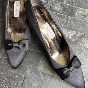 May include: A pair of black satin pumps with pointed toes and small bows. The shoes have a low heel and the Calvin Klein label is visible inside. The shoes are displayed on a textured surface.