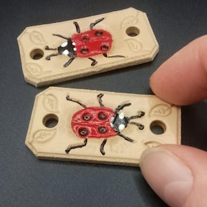 May include: Two rectangular tan leather tags featuring a hand-painted red ladybird with black spots and white accents. Each tag has two circular holes on the ends and embossed leaf patterns on the corners, providing a functional design for attachment.