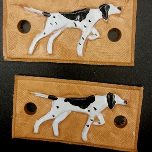 May include: Two rectangular tan leather patches, each featuring a raised, hand-painted white dog with black spots and a black head. Each patch has two circular holes punched on the left and right sides for attachment to a garment or accessory.