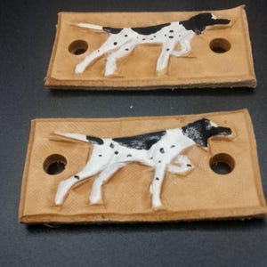May include: Two rectangular tan leather tags, each featuring a raised, hand-painted white and black spotted dog in a running pose. Each tag has two circular holes punched on the left and right sides for attachment, with a textured surface.