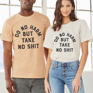 Puede incluir: Dos personas con camisetas a juego con el texto "Do No Harm But Take No Shit" impreso en un semicírculo negro en la parte delantera. La persona de la izquierda lleva una camiseta marrón claro y la persona de la derecha lleva una camiseta blanca.