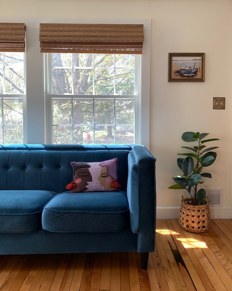 K&ouml;nnte beinhalten: Ein blaues Samtsofa mit einem Kissen, das eine farbenfrohe Illustration von zwei Gesichtern zeigt. Das Sofa steht vor einem Fenster mit Blick auf B&auml;ume und ein Haus. Rechts im Bild steht eine Topfpflanze.