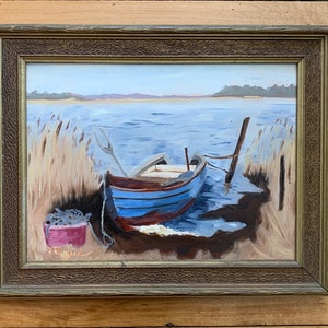 K&ouml;nnte beinhalten: Ein gerahmtes Gem&auml;lde, das ein blaues und braunes Boot zeigt, das an einem Seeufer angelegt ist. Das Boot ist an einem Holzpfosten festgemacht, und im Vordergrund ist ein Eimer mit Fisch zu sehen. Das Gem&auml;lde ist unten links mit "E. Girard" signiert.