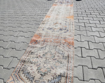 May include: A rectangular, distressed-look rug with a faded, vintage aesthetic. The rug features a central design with geometric patterns in shades of orange, blue, and beige. The rug is placed on a gray, textured surface.