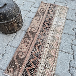 Op de afbeelding: Een rechthoekig geweven kleed met een geometrisch patroon in tinten bruin, zwart en gedempt roze, geplaatst naast een donkere, verweerde metalen container met een afgerond deksel, een handvat en zichtbare klinknagels op het buitenoppervlak.