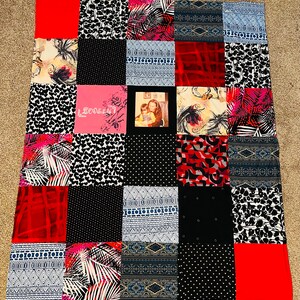 May include: A patchwork quilt with a variety of patterns and colors, including red, black, white, and blue. The quilt features a small photo of a mother and child in the center. The quilt is made of fabric scraps and has a handmade feel.