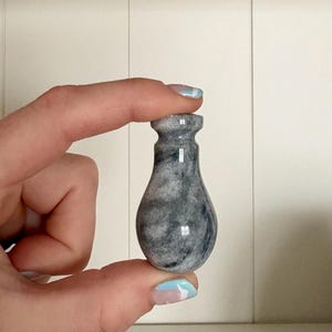 May include: A small, gray and white marble vase with a bulbous body and a narrow neck. The vase is held between two fingers. The vase is approximately 3 inches tall. The marble has a swirling pattern.