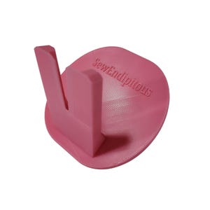 May include: A pink sewing thread spool holder with a curved base and a vertical support. The text "SewEndipitous" is printed on the base. The holder is designed to keep thread spools organized.