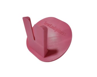 May include: A pink sewing thread spool holder with a curved base and a vertical support. The text "SewEndipitous" is printed on the base. The holder is designed to keep thread spools organized.