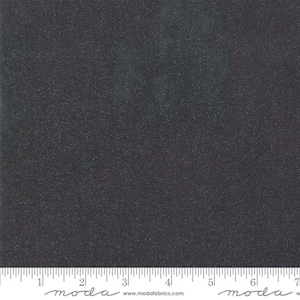 May include: Dark gray fabric with a subtle, speckled pattern. The fabric has a textured appearance, with tiny, light-colored dots scattered across the surface. A ruler is at the bottom, with the word "moda" printed on it.