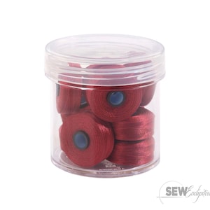 May include: A clear plastic jar filled with several spools of red thread. Each spool has a blue center. The jar has a clear lid and is sitting on a white surface. The thread is tightly wound.