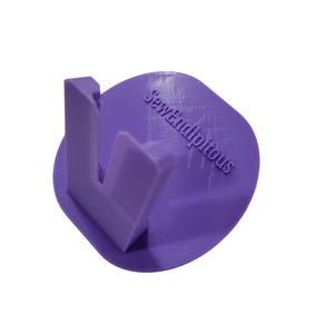 May include: A lavender-colored 3D-printed sewing thread holder. The holder has a curved base with the text "SewEndipitous" and an L-shaped thread guide. The design is simple and functional, ideal for organizing sewing supplies.