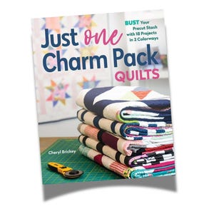 May include: Book cover with the title "Just one Charm Pack Quilts" and the subtitle "Bust Your Precut Stash with 18 Projects in 2 Colorways". The cover features a stack of colorful quilts and a rotary cutter on a green cutting mat.