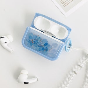 May include: A light blue protective case for wireless earbuds with a floral design. The case has a white strap with silver accents and a small silver charm.