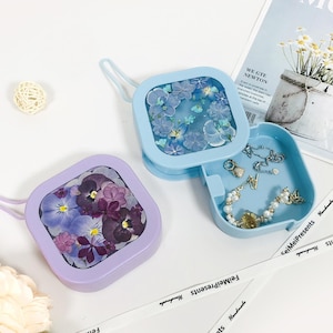 May include: Two square-shaped, hinged boxes with a clear window on the top. The boxes are light blue and lavender and feature pressed flowers inside. The boxes are open and the light blue box contains a small collection of jewelry.