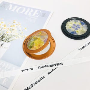 May include: Two small, round, handheld mirrors with a floral design. The mirror on the left has a gold frame and a yellow and white floral design. The mirror on the right has a black frame and a blue and white floral design. Both mirrors have a white ribbon with the text "FeiMeiPresents Handmade" printed on them.