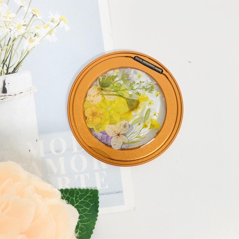 May include: A round, gold-colored compact mirror with a clear lid. The lid features a pressed flower design with yellow, purple, and white flowers.