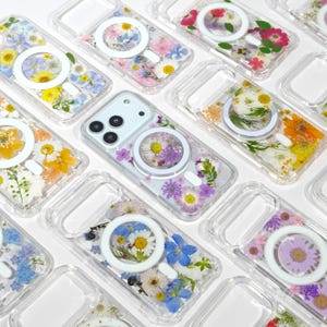 May include: Clear phone cases with embedded dried flowers in various colors, including yellow, pink, and purple. Each case has a white ring for a magnetic charger. The cases are arranged on a white surface.