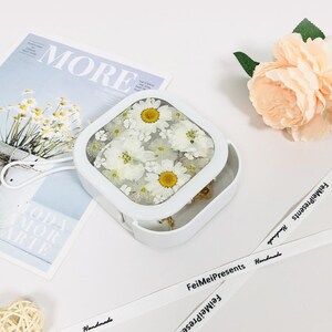 Könnte beinhalten: Weiße quadratische Box mit einem durchsichtigen Deckel, in der gepresste weiße Blumen zu sehen sind. Die Box steht auf einer weißen Oberfläche mit einem weißen Band mit schwarzem Text, der "FeiMeiPresents Handmade" lautet.