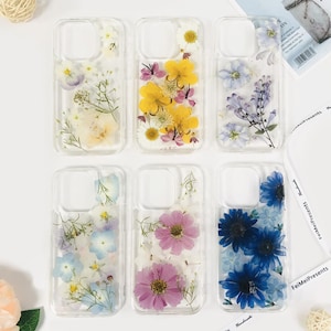 Può includere: Sei custodie trasparenti per telefono con fiori pressati. Le custodie presentano varie composizioni floreali, tra cui fiori gialli, viola e blu. Le custodie sono disposte su una superficie bianca.