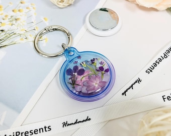 Puede incluir: Un llavero redondo transparente con un borde azul y morado, lleno de flores moradas y rosas secas. El llavero tiene un cierre de anillo plateado. Se ve una cinta blanca con la palabra "Handmade".