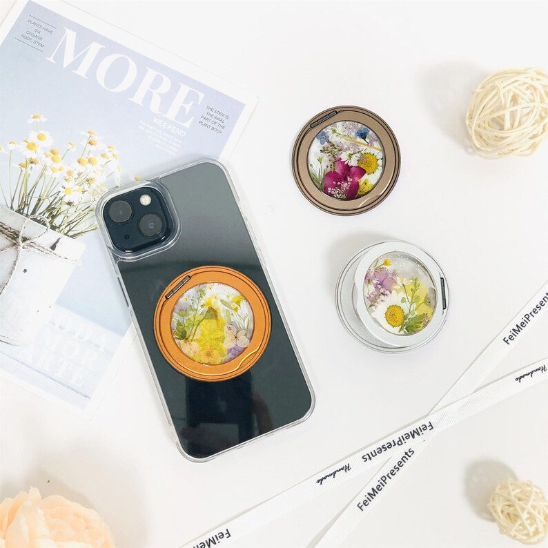 May include: A clear phone case with a black background and a gold ring holder with pressed flowers inside. The ring holder is attached to the back of the phone case. There are three ring holders in the image, each with a different color and flower design.