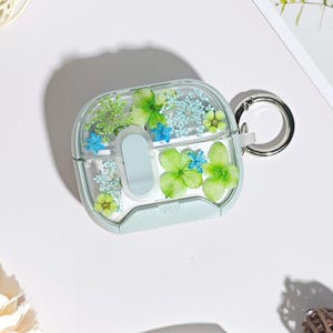 Puede incluir: Funda transparente para AirPod con marco verde menta, llena de flores secas verdes y azules. La funda tiene un anillo de metal plateado y la palabra "OPEN" es visible en la parte inferior.