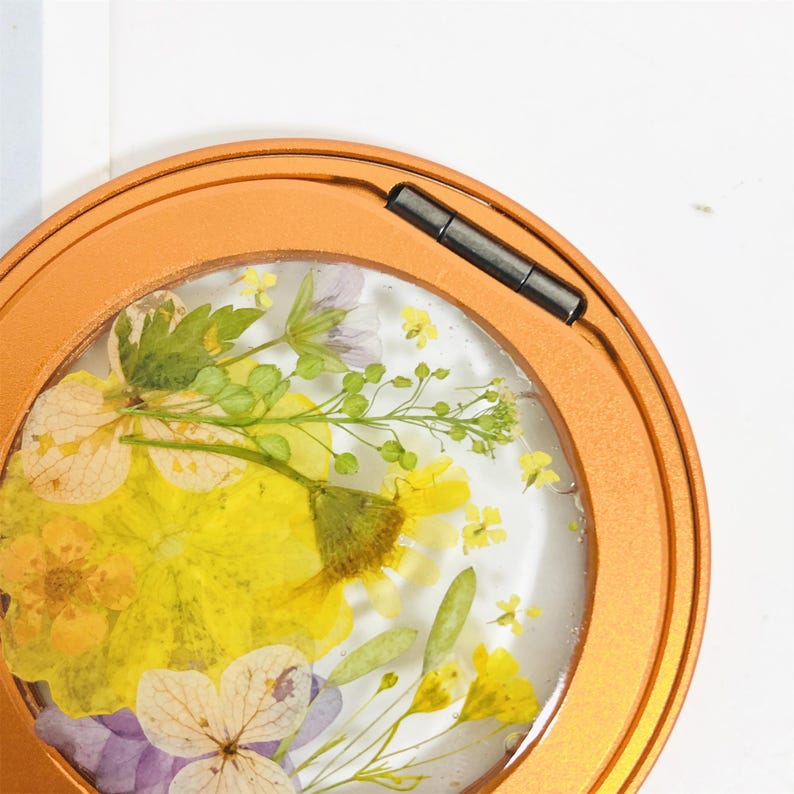 May include: A round, gold-colored metal frame with a clear glass insert containing pressed flowers. The flowers are yellow, white, and purple.