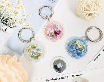May include: Keychains with dried flowers encased in clear resin, attached to silver-colored metal rings. One keychain has pink flowers, another has blue flowers, and a third has white and blue flowers. An Apple AirTag is also visible.