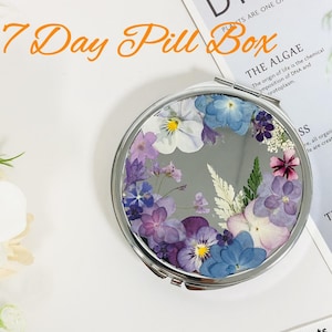 May include: A silver compact mirror with a round, clear glass lid. The lid is decorated with pressed flowers in shades of purple, blue, and white. The mirror is labeled "7 Day Pill Box".