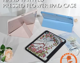 Op de afbeelding: Handgeschilderde iPad-hoesjes in roze en blauw, met een design van geperste bloemen. Eén hoesje is open, met de tablet zichtbaar, terwijl een ander is opgevouwen tot een standaard. De tekst luidt "HAND-PAINTED PRESSED FLOWER IPAD CASE."