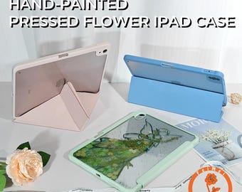 Op de afbeelding: Handgeschilderde iPad-hoesjes met geperste bloemen in pastelkleuren. Eén hoesje is lichtroze, een ander lichtblauw en een derde lichtgroen met een ontwerp van geperste bloemen. De hoesjes zijn open en tonen de iPad-schermen.
