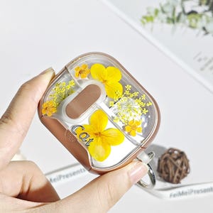 Può includere: Una custodia rettangolare trasparente per AirPods con una cornice color oro, con fiori gialli e la parola "Cat". La custodia ha un interno marrone e un attacco ad anello argentato. La custodia è tenuta in mano.