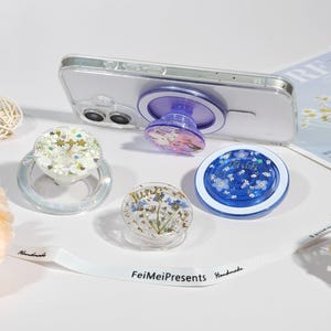 Op de afbeelding: Een verzameling decoratieve telefoonhouders in verschillende ontwerpen. Eén is bevestigd aan een doorzichtige telefoonhoes. De houders hebben bloemen- en glitterelementen in helder, blauw en wit. De houders zijn rond en hebben een pop-out functie.