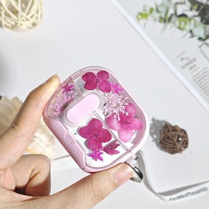 Può includere: Una custodia rosa e trasparente per auricolari wireless, decorata con fiori rosa pressati. La custodia ha un accento rosa chiaro e un anello argentato per attaccare un cordino. La parola "OPEN" è visibile sulla custodia.
