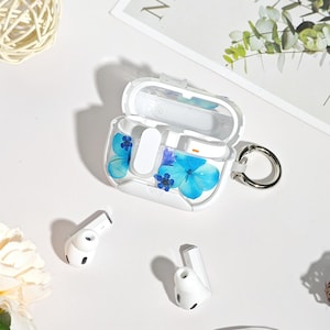 Può includere: Una custodia bianca per AirPods Pro con coperchio trasparente, decorata con fiori blu pressati. La custodia è aperta, rivelando gli auricolari bianchi. Un moschettone argentato è attaccato.