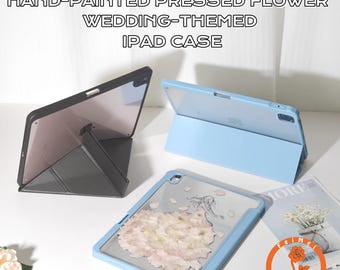 Op de afbeelding: Handgeschilderde iPad-hoesjes met geperste bloemen met een bruiloftthema in zwart en lichtblauw. Eén hoesje heeft een ontwerp met geperste bloemen, een ander is effen lichtblauw en de derde is zwart. De hoesjes zijn open en tonen de iPad-schermen.