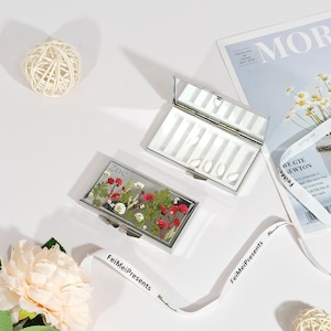 Puede incluir: Dos pastilleros rectangulares plateados con espejos. Uno está abierto, revelando compartimentos blancos. El otro presenta un diseño floral con flores rojas y blancas y el nombre "Zoe". También se ve una cinta blanca con "FeiMeiPresents".