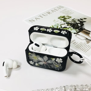 Op de afbeelding: Zwarte Airpods-hoes met een bloemenpatroon en een wit parelkoord. De hoes is open en de Airpods zitten erin. De hoes heeft een kleine lus aan de zijkant.