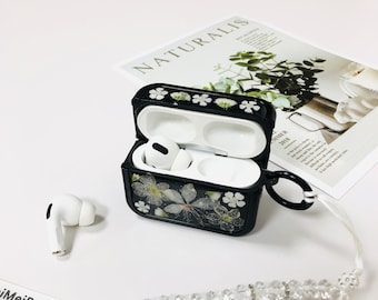 May include: Black Airpods case with a floral design and a white beaded strap. The case is open and the Airpods are inside. The case has a small loop on the side.