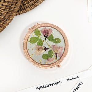 Könnte beinhalten: Ein runder, roségoldener Kompakt-Spiegel mit einem Design aus gepressten Blumen. Der Spiegel hat einen weißen Hintergrund mit rosa Blumen und grünen Blättern. Die Blumen sind gepresst und in Harz konserviert. Der Spiegel ist von einem roségoldenen Metallrahmen umgeben.
